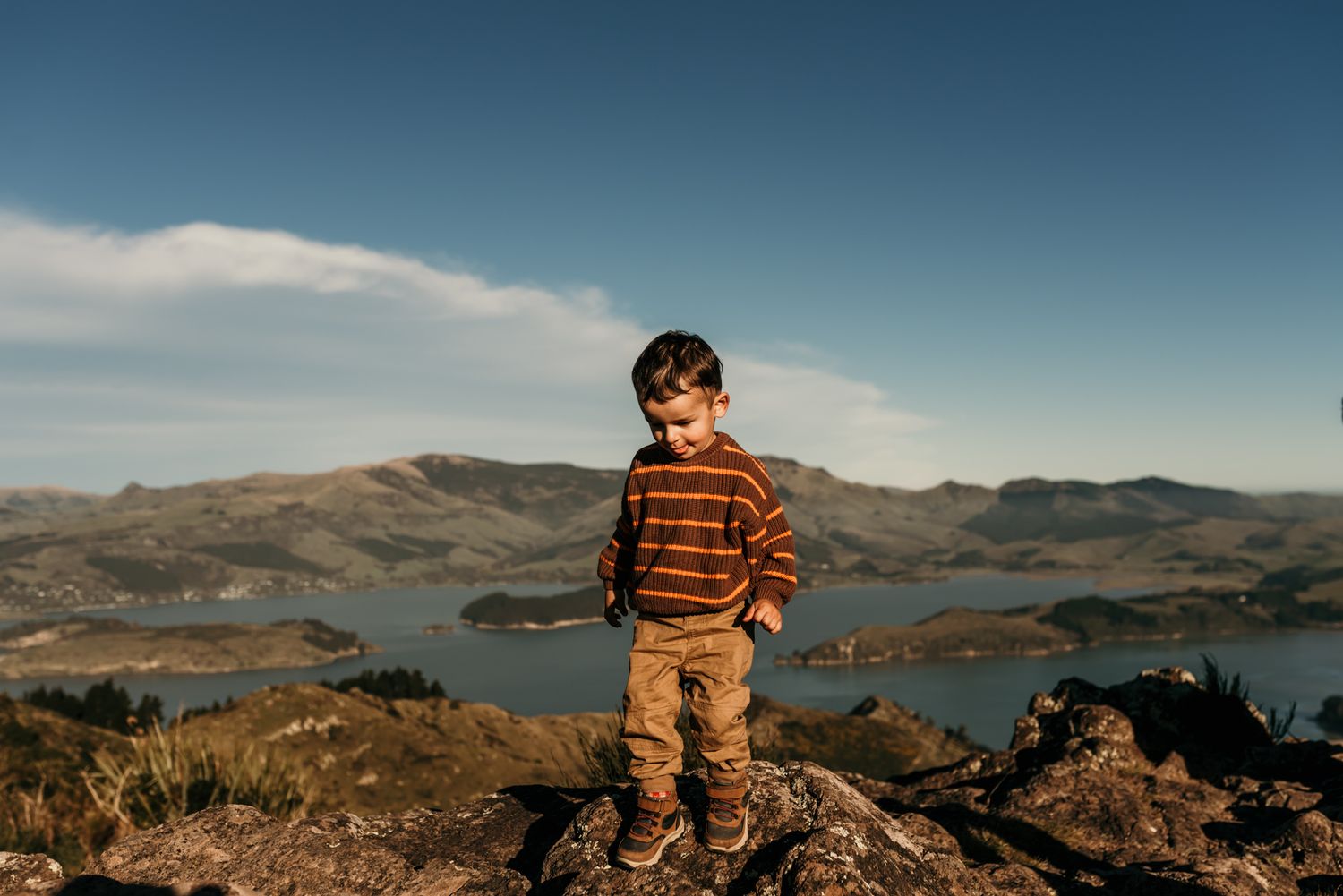 Child in orange striped sweater stands confidently on rocky overlook with lake and mountains in background.