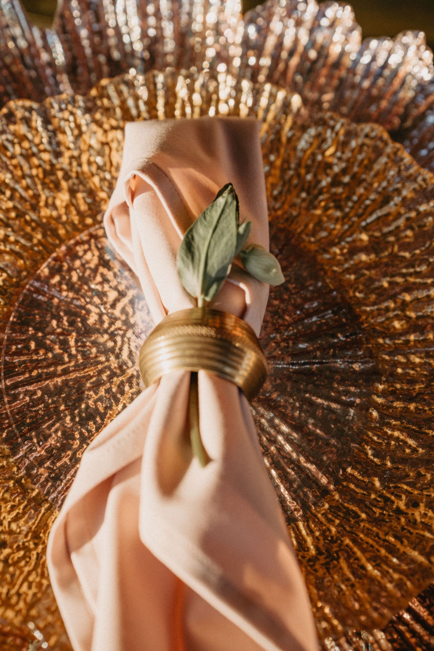 Pink silk napkin with gold ring and olive leaf on decorative gold charger plate.
