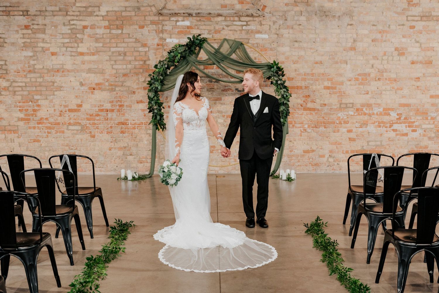 Bride and groom holding hands looking at eachother smilling in front of allter and brick wall.