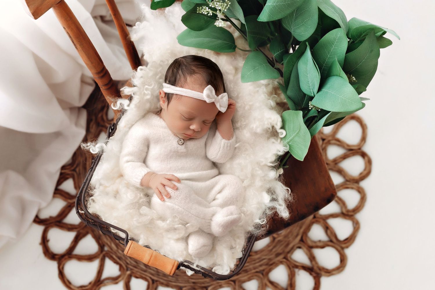 Baby in white clothing rests in decorative brown wicker basket with green plants.