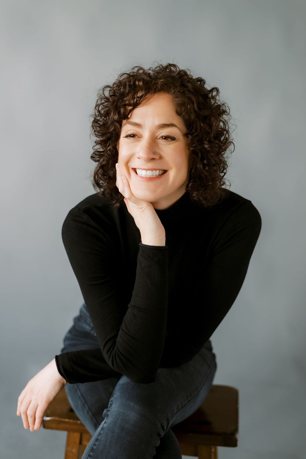 Professional headshot of person in black turtleneck with curly brown hair smiling warmly against gray background.