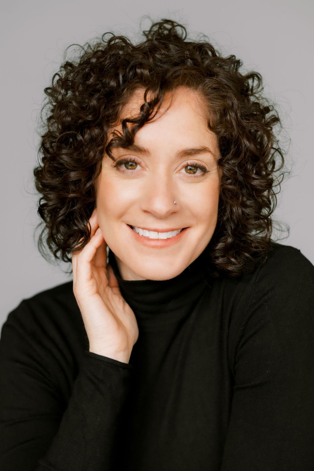 Close-up portrait with natural curly hair and black turtleneck against neutral background.