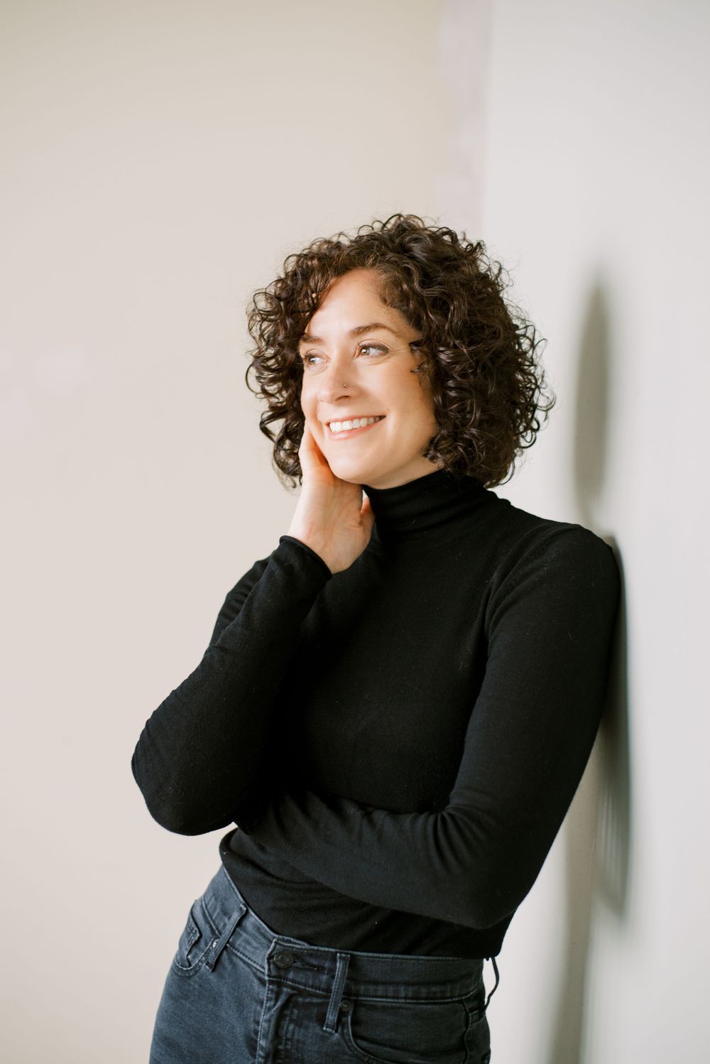 Casual portrait against white wall wearing black turtleneck and jeans with natural curly hair.