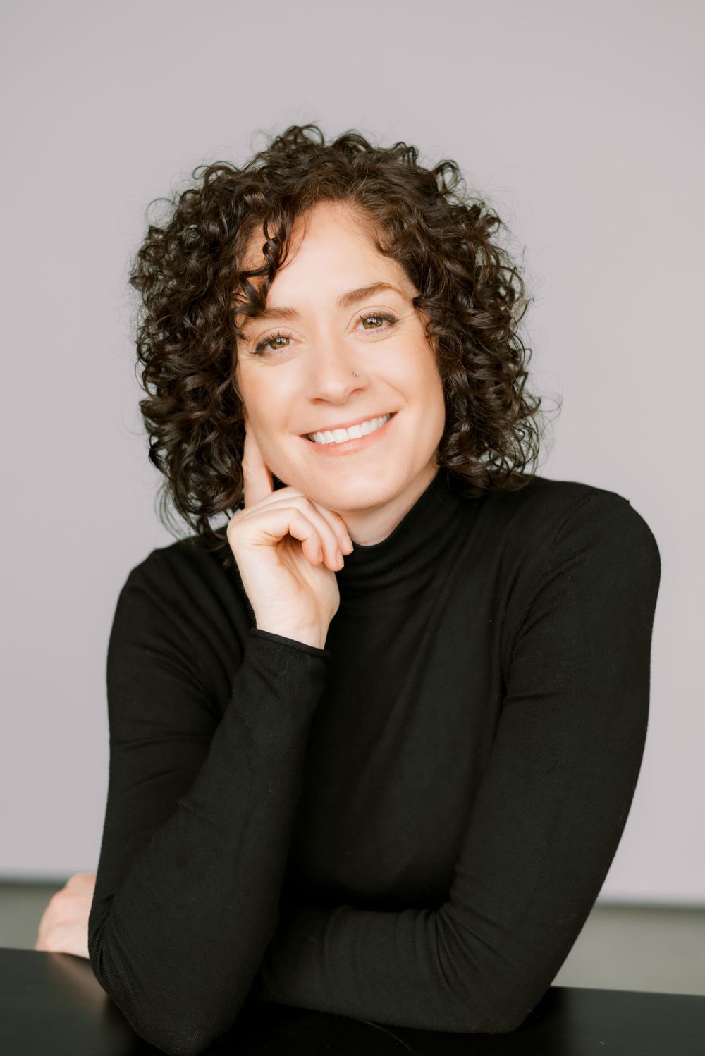 Professional headshot with natural curly hair and black turtleneck against simple background.