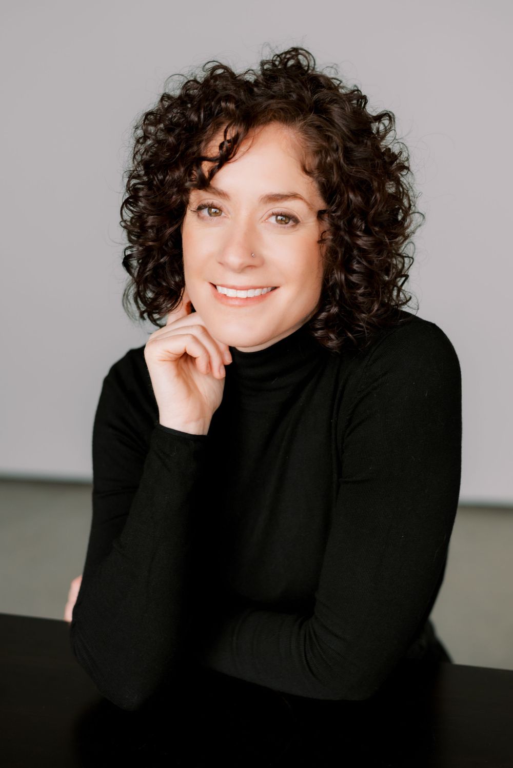 Portrait of person in black turtleneck with natural curly hair and bright smile against gray backdrop.