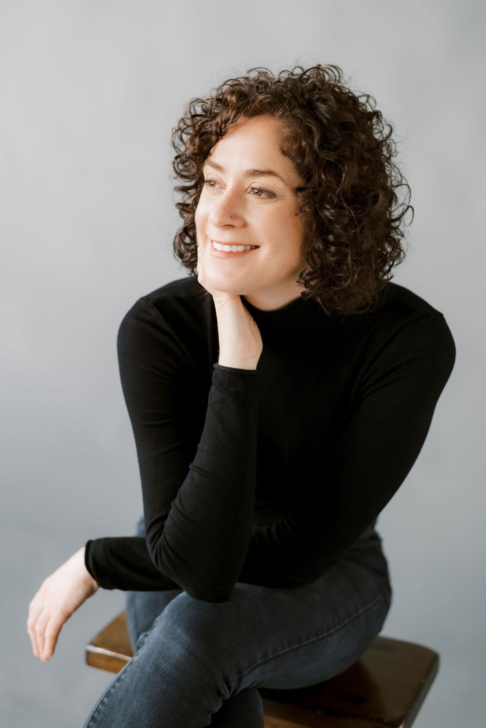 Professional portrait of someone wearing a black turtleneck sweater and jeans while seated with curly brown hair.