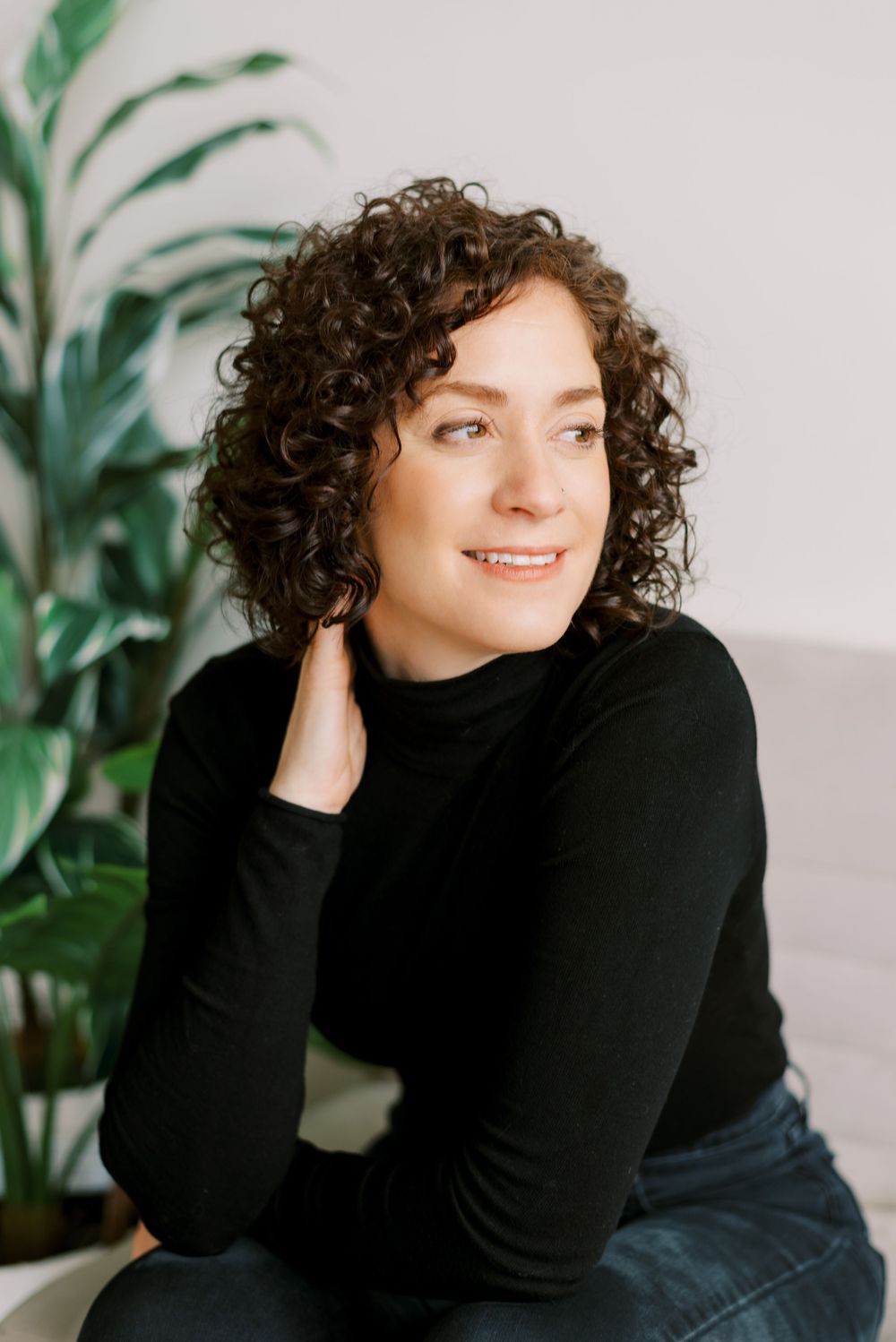 Natural light portrait with curly hair in black sweater beside decorative plant.