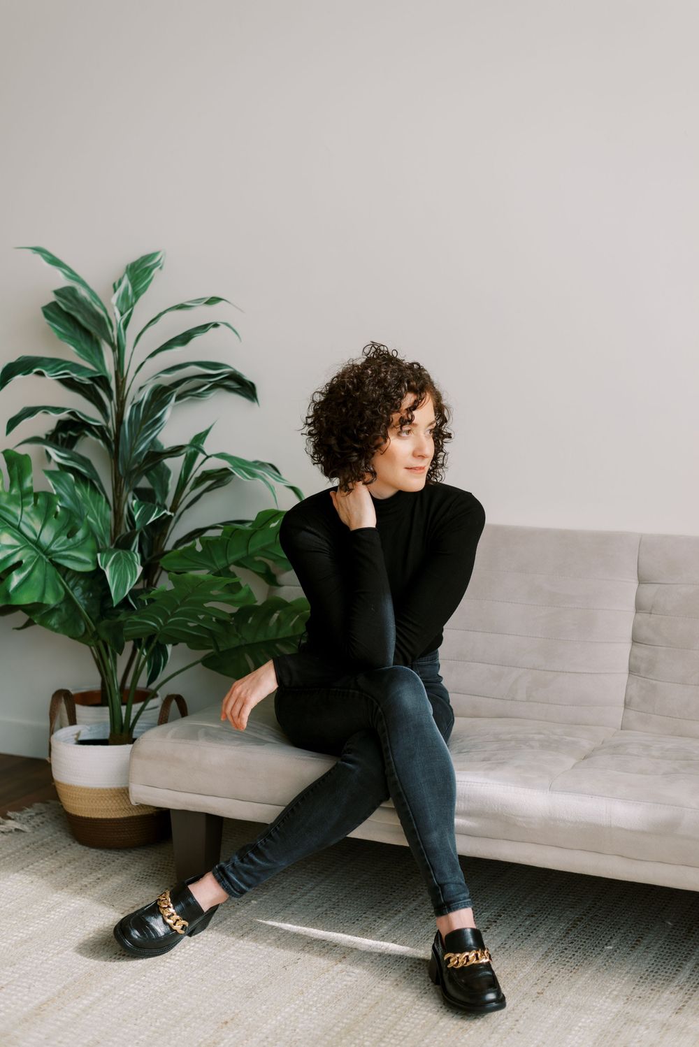 Lifestyle portrait on grey couch with potted plant wearing black sweater and jeans.