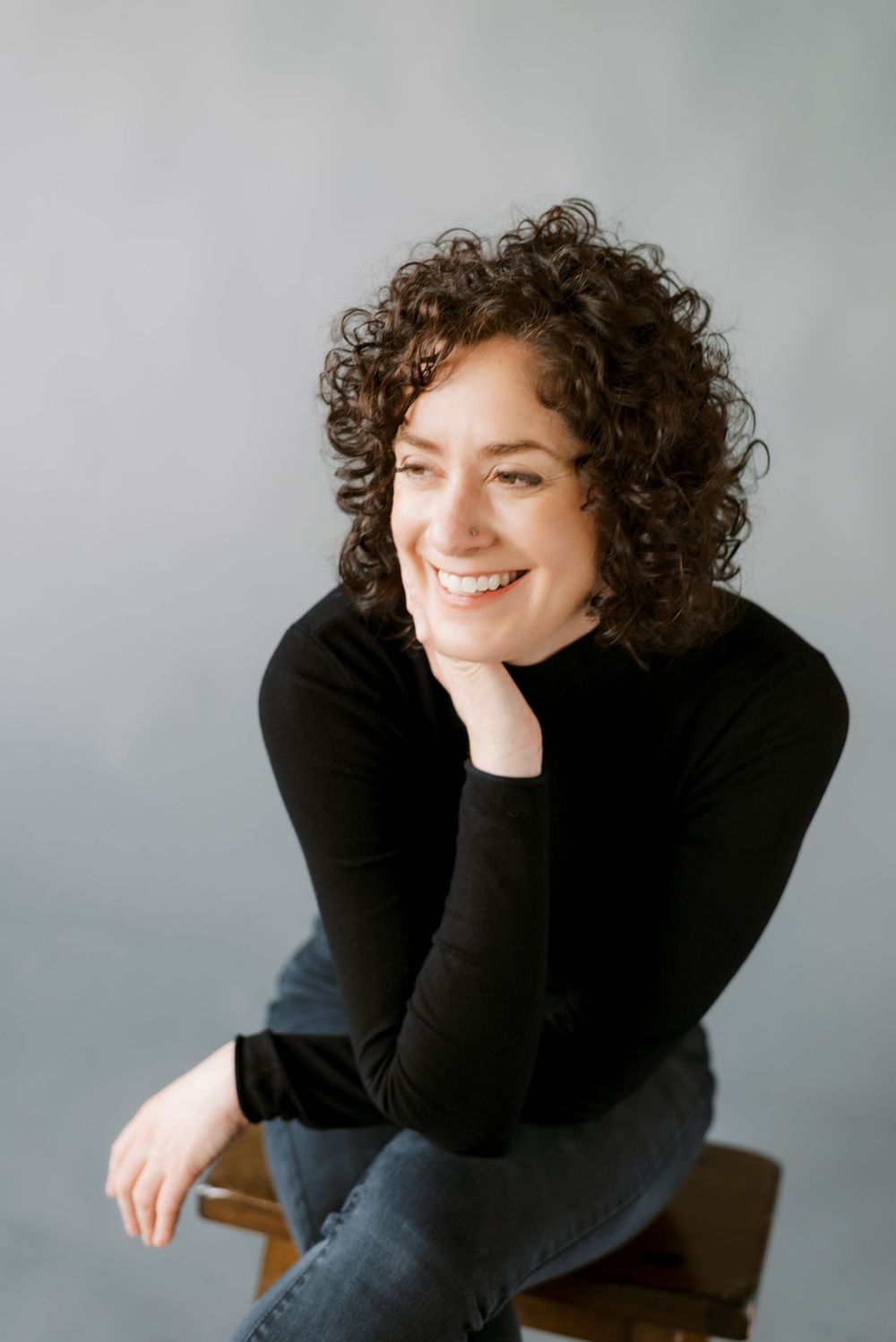 Portrait with natural curly hair in black turtleneck smiling warmly against grey background.