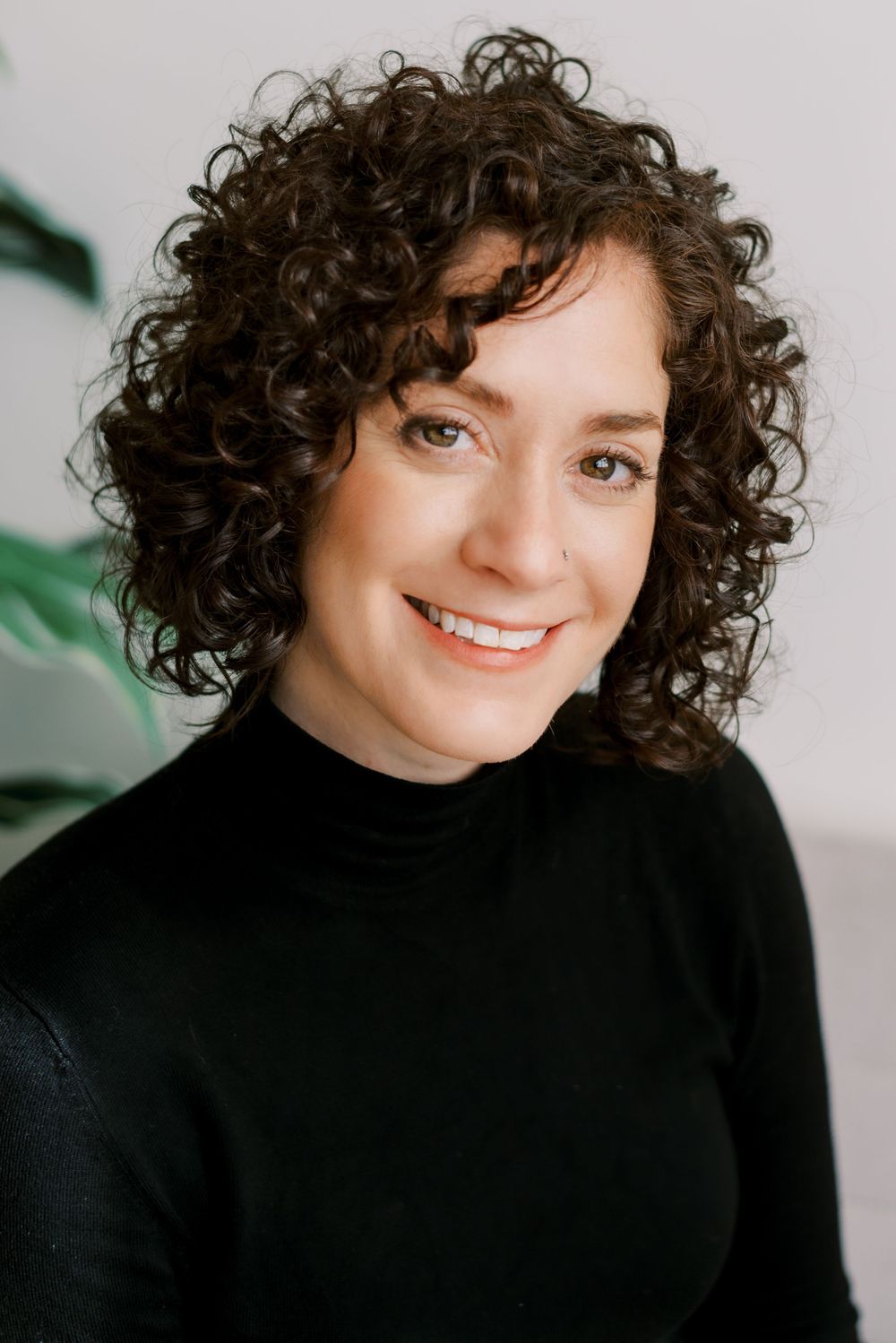 Professional headshot with curly hair and black turtleneck near indoor plant.