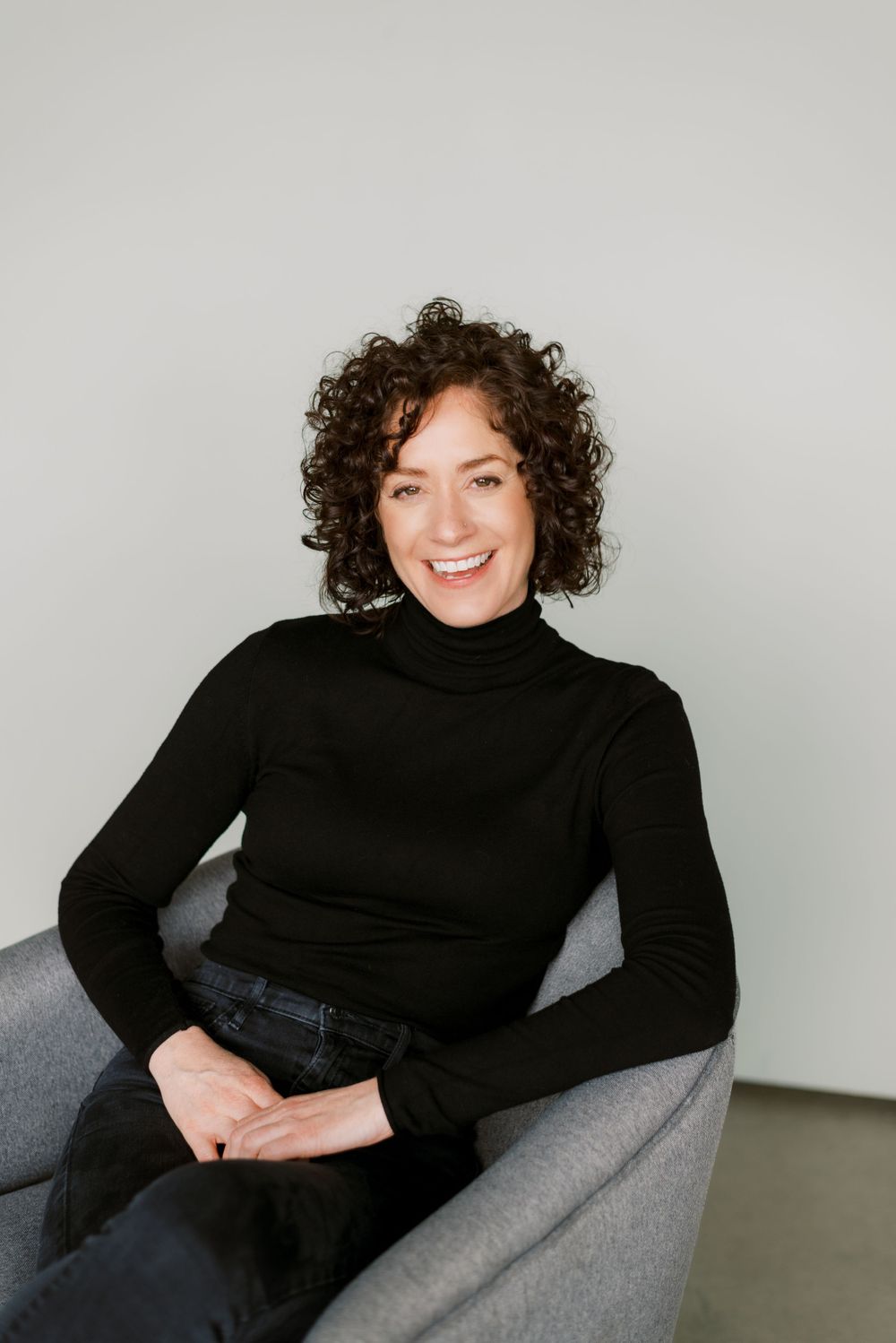 Person with curly brown hair sits relaxed in gray chair wearing black turtleneck and jeans.