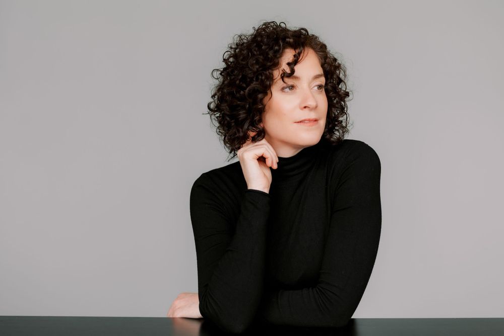 Minimalist studio portrait in black turtleneck with curly hair against grey background.