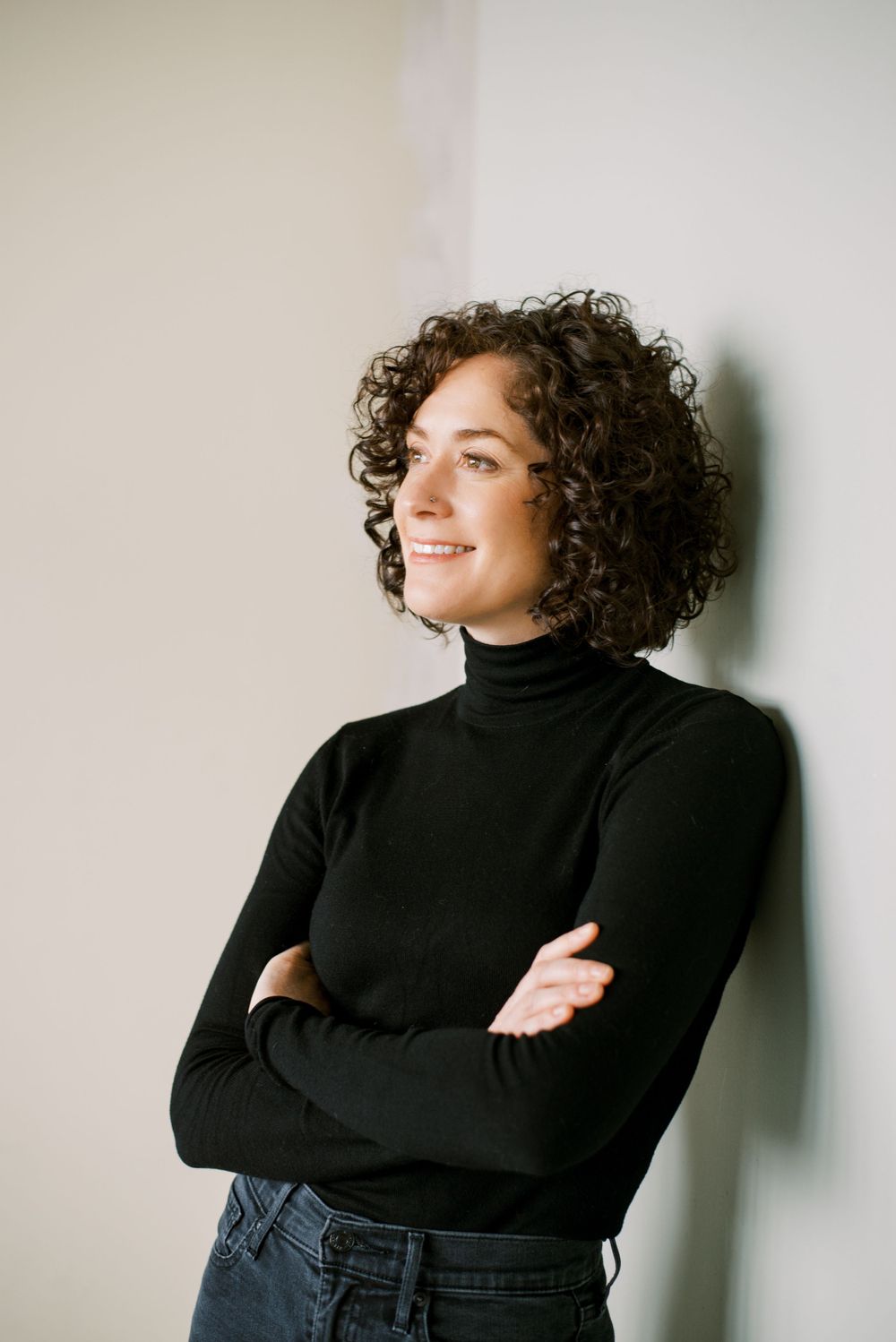 Studio portrait showing confident pose in black turtleneck with arms crossed and curly hair.