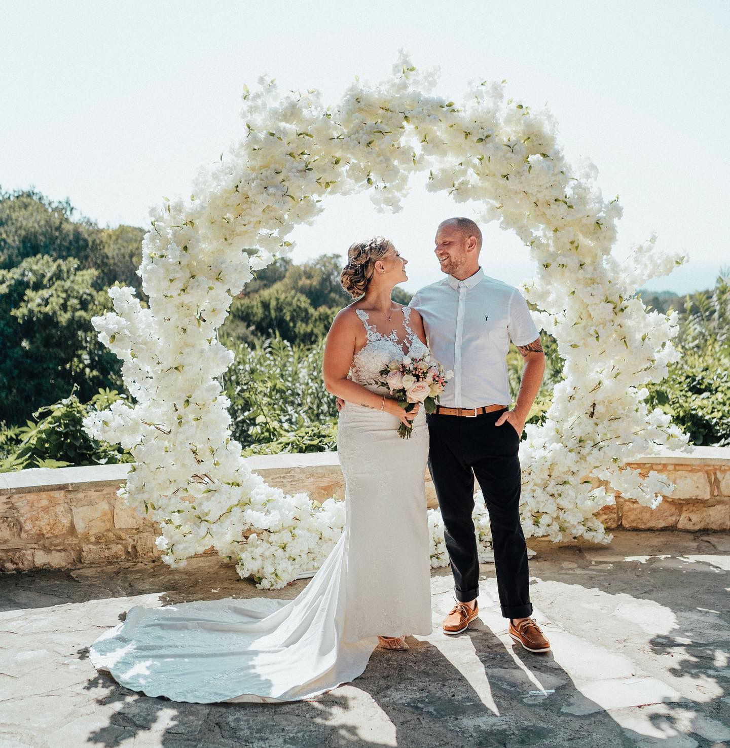 Wedding couple standing under ornate white floral arch in bright sunlight.
