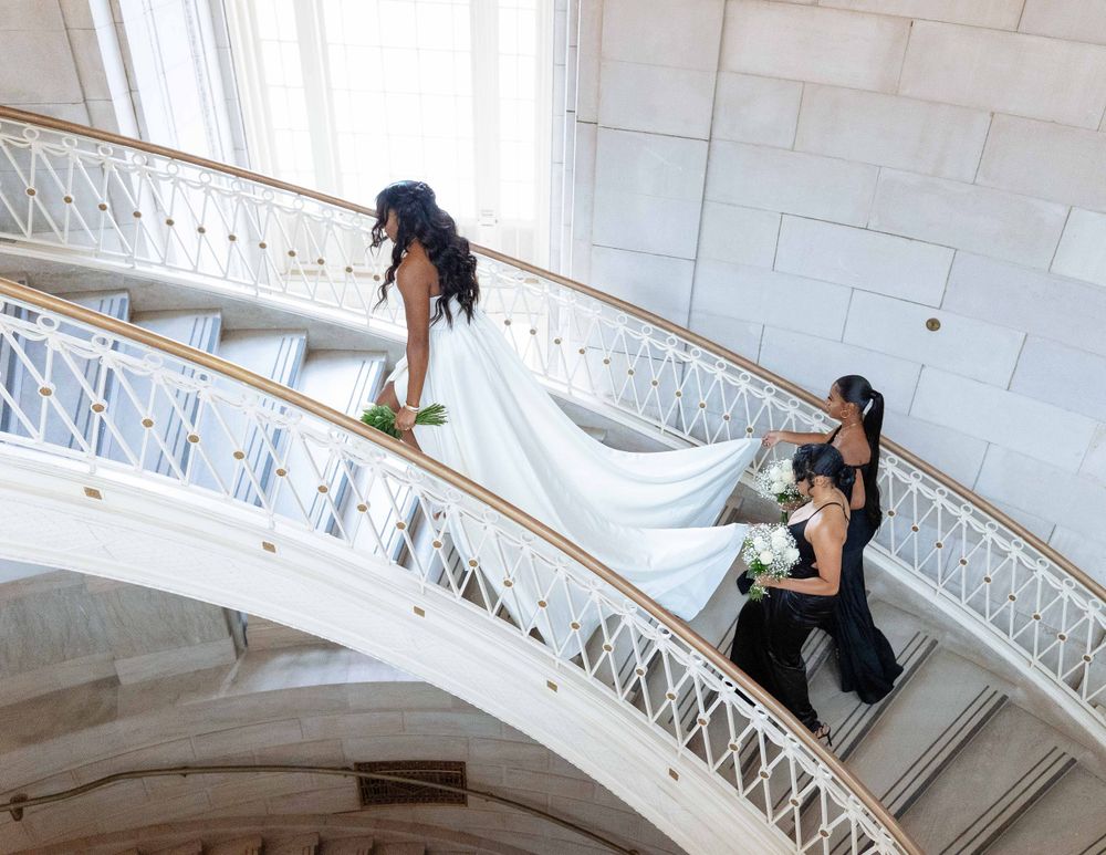 Two people in wedding attire descend a grand marble spiral staircase.