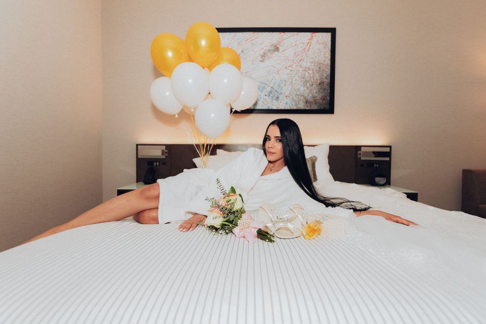 A newlywed relaxes on a white bed with yellow and white balloons and flowers as decorations.