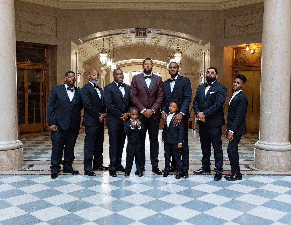 A formal wedding party in matching tuxedos poses in an elegant marble hallway.