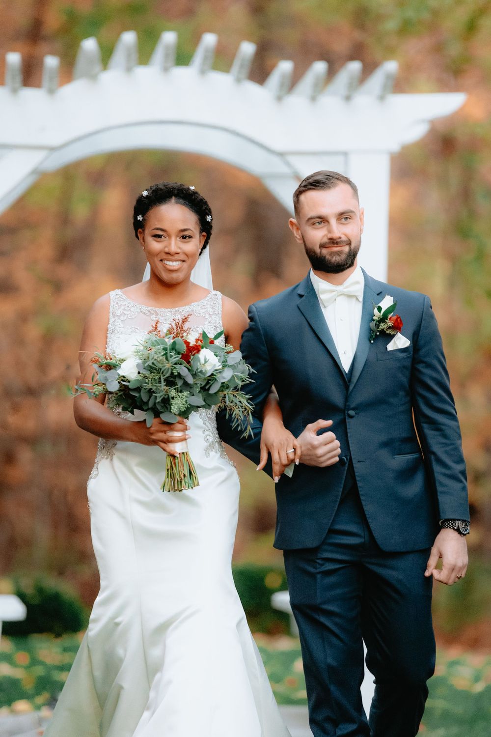 A couple poses under a white garden arch during their autumn outdoor wedding ceremony.