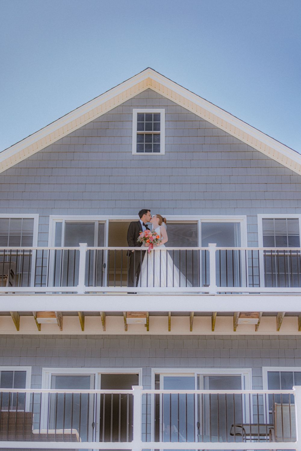 Wedding couple embracing on white beach house balcony against blue sky.