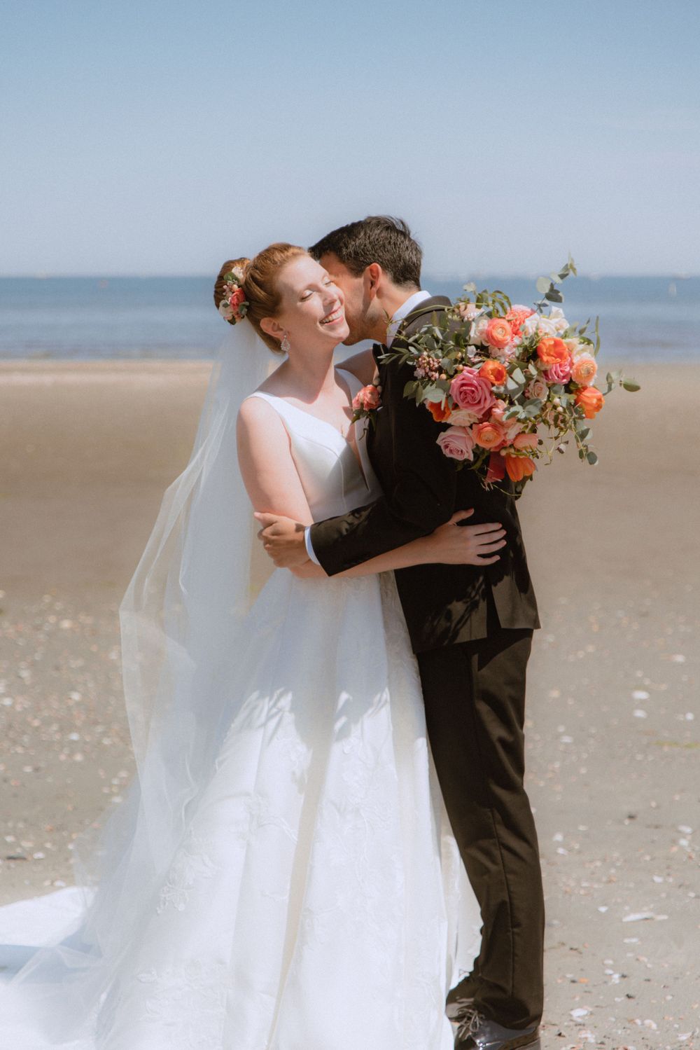 Joyful wedding moment captured on sunny beach with coral floral bouquet.