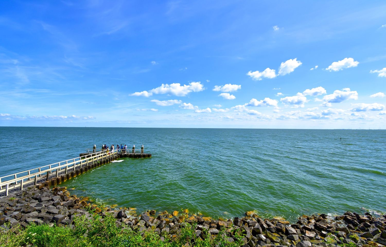A wooden pier extends into turquoise waters under bright blue skies with white fluffy clouds.