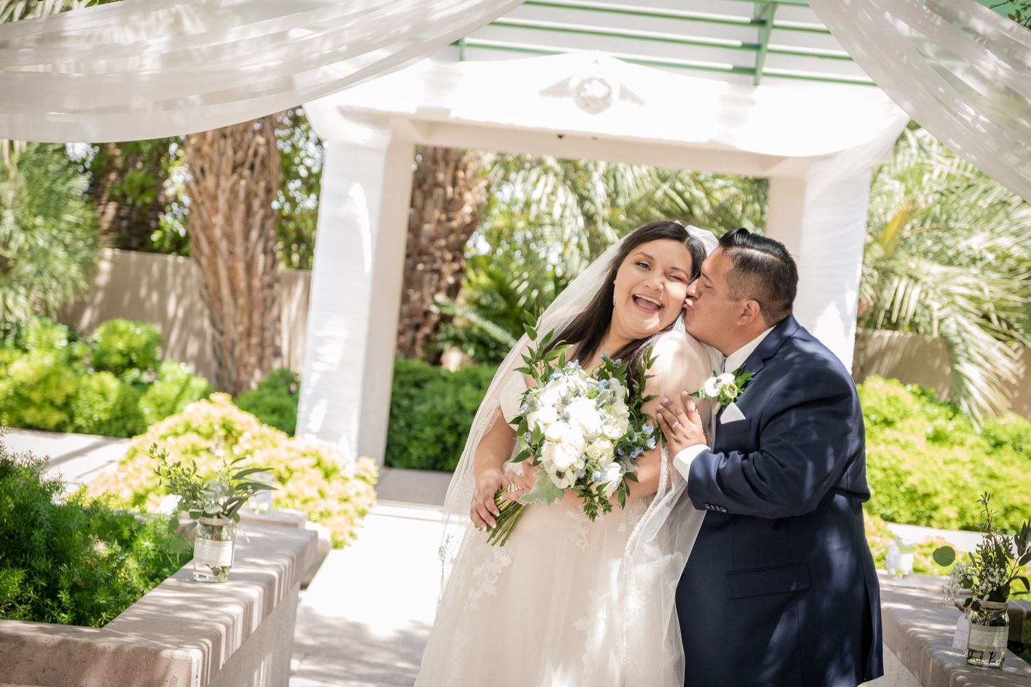 Sunny outdoor wedding portrait in a garden gazebo with lush greenery.
