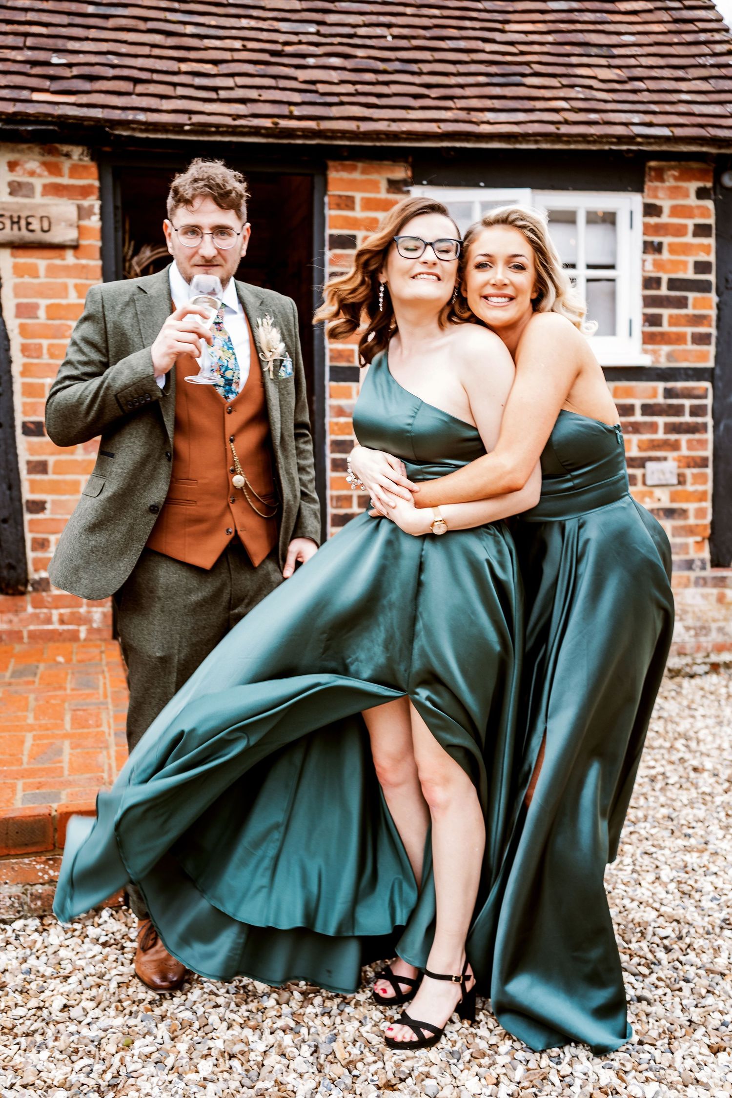 Wedding party members in emerald gowns pose playfully outside a brick building.