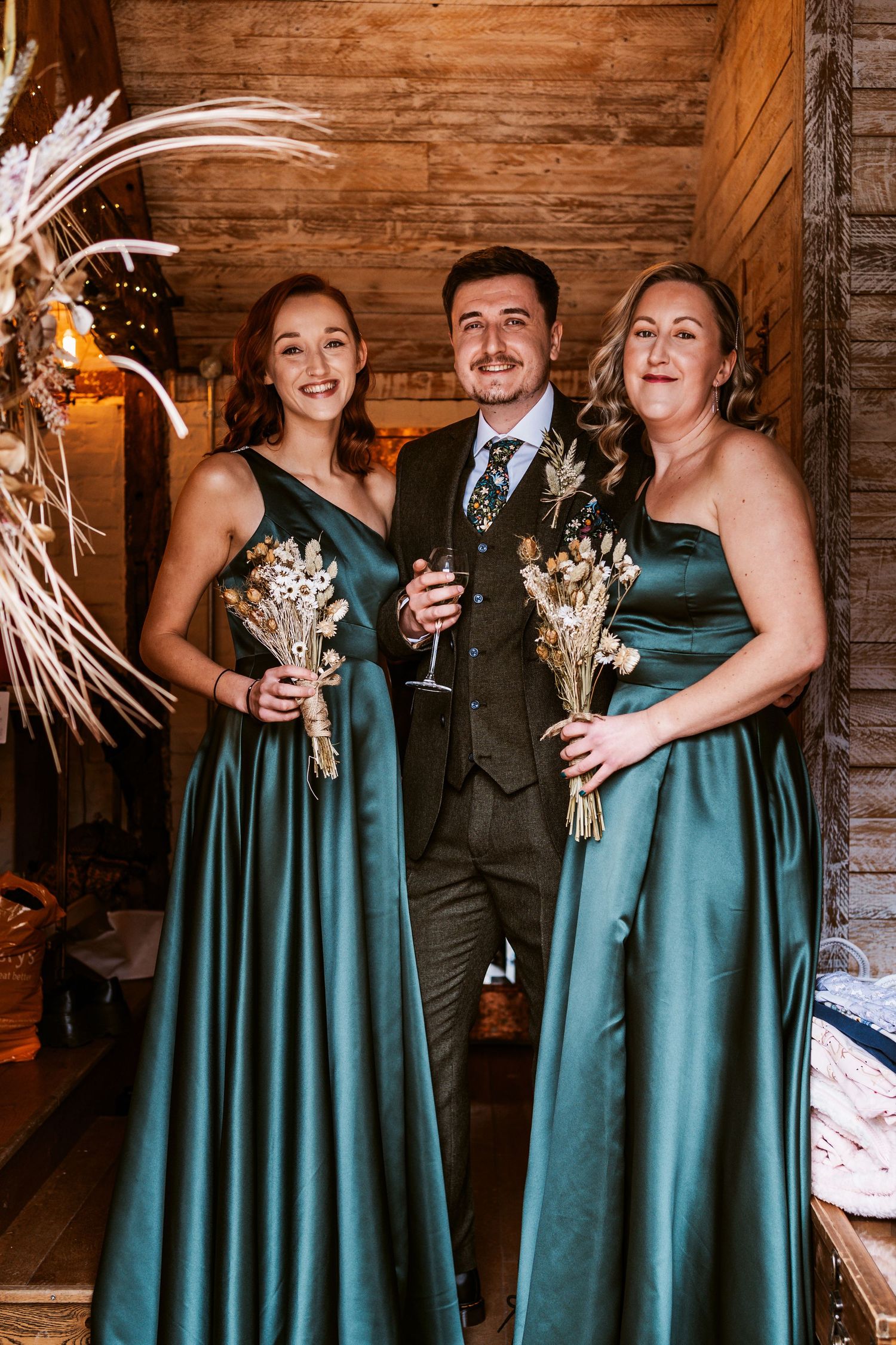 Three wedding attendees in emerald dresses pose with bouquets in a rustic barn setting.