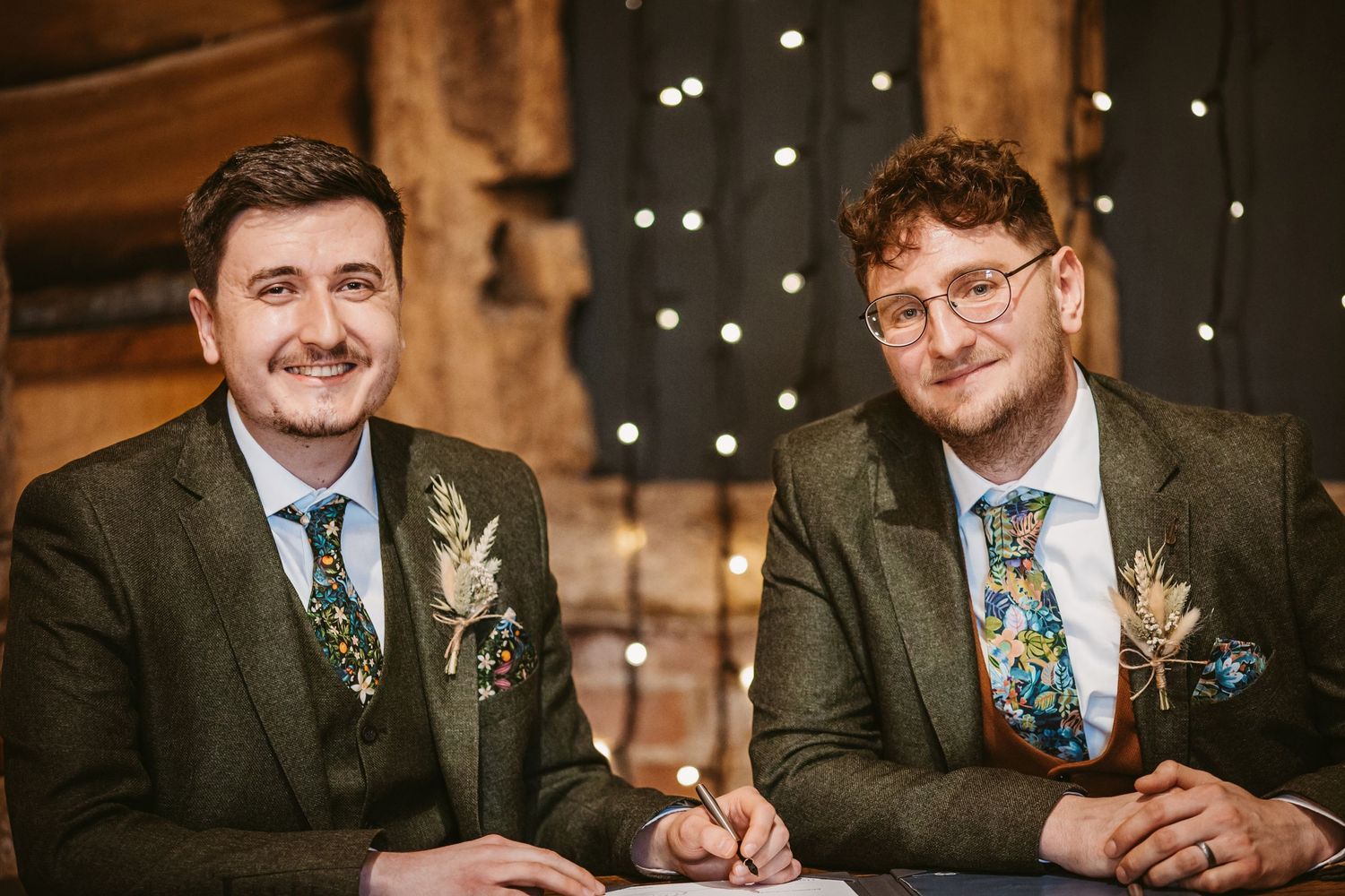 Wedding couple smiling while seated at rustic wooden table decorated with floral boutonnieres.