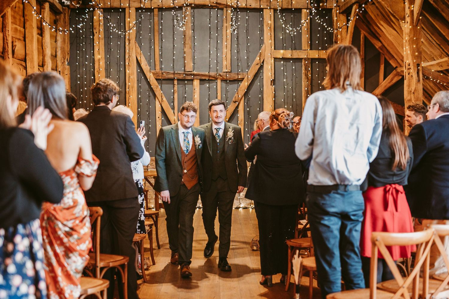 Wedding guests stand and applaud during ceremony in rustic barn setting with string lights.