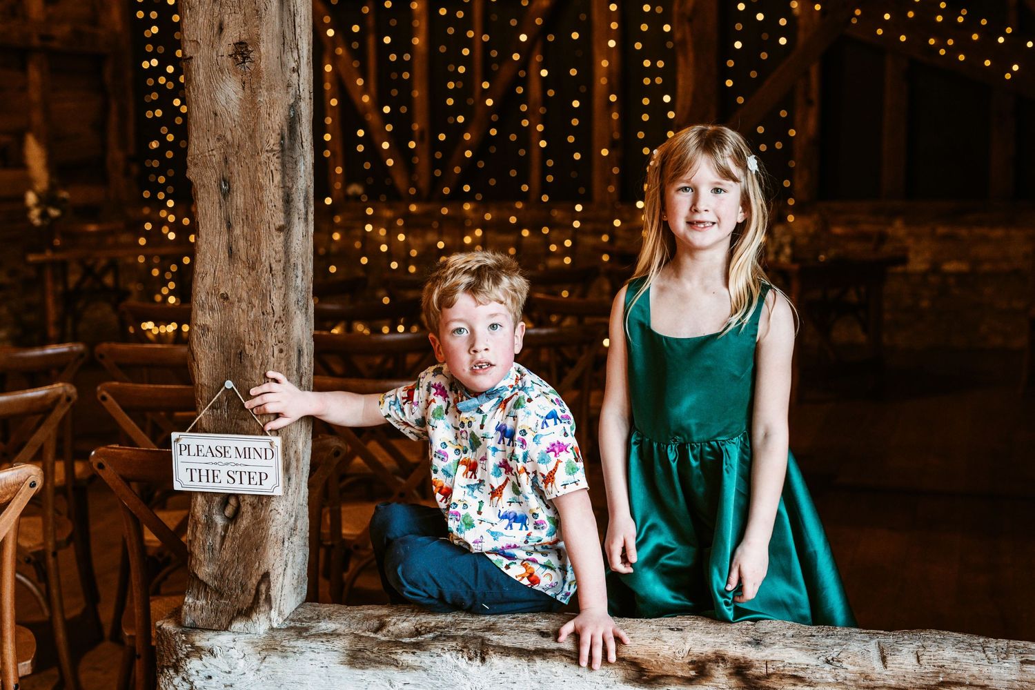 Children sitting on rustic wooden ledge inside barn decorated with twinkling lights.