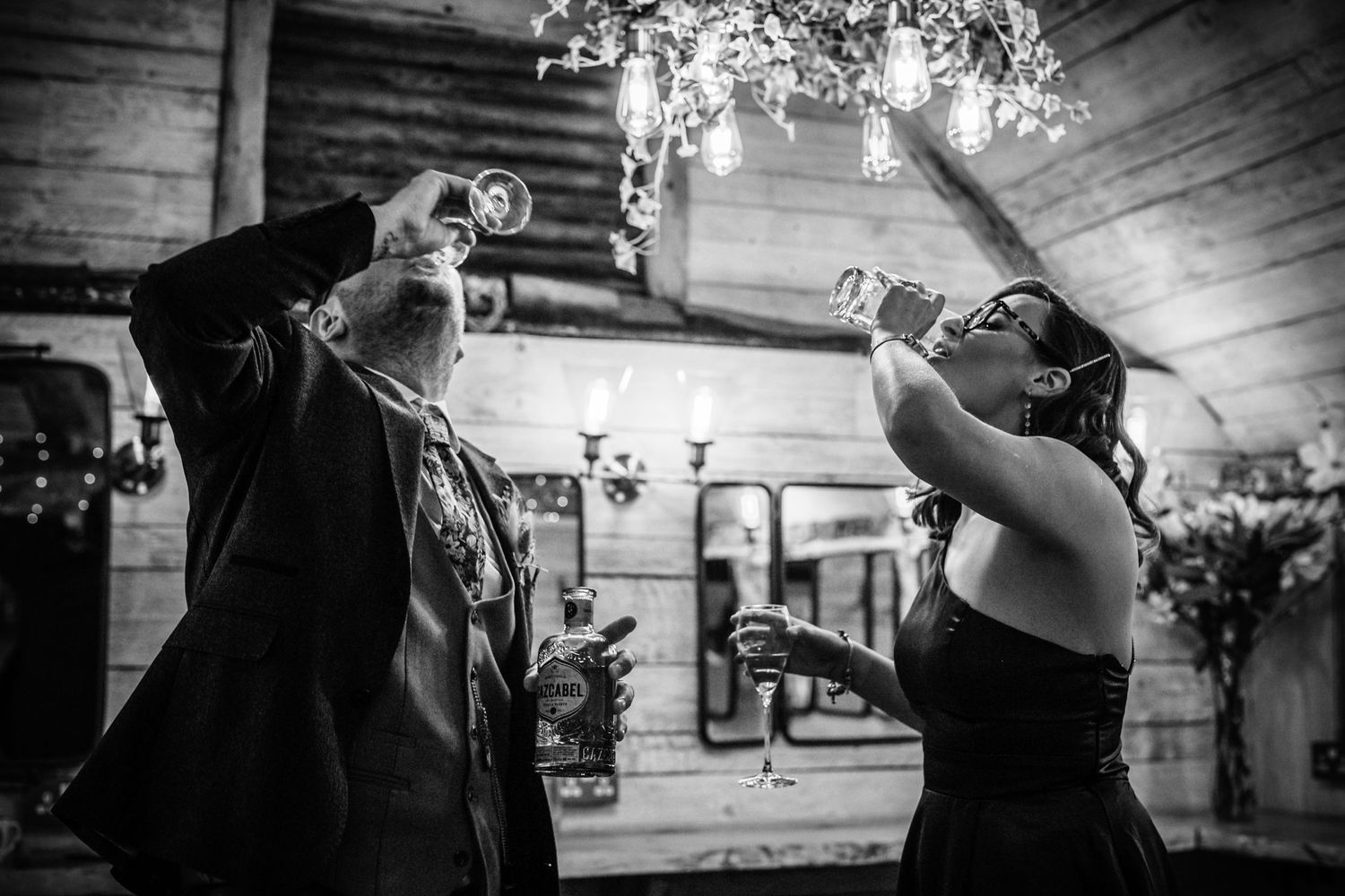 Black and white photo of wedding guests toasting and drinking from champagne bottles in rustic venue.