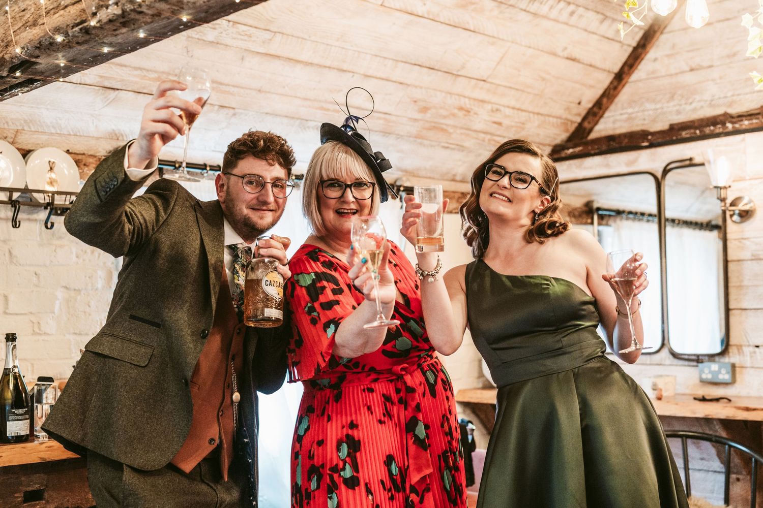 Wedding guests in vintage attire celebrating with drinks in rustic indoor venue.