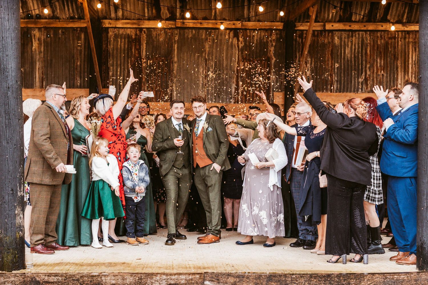Large wedding party celebration with guests throwing confetti outside rustic barn venue.
