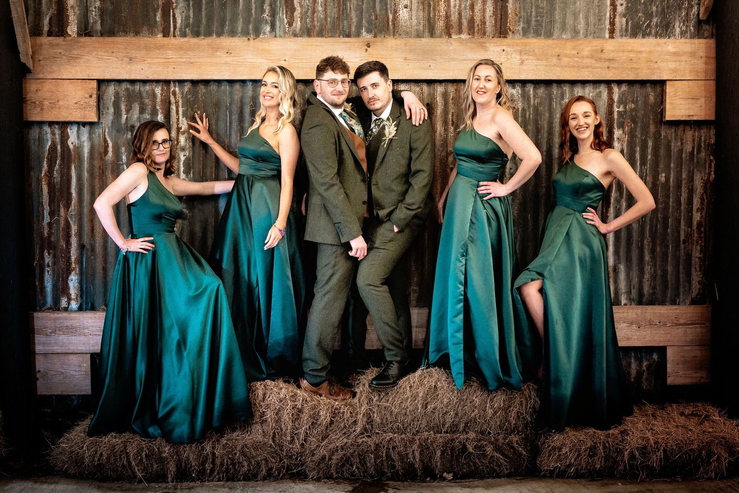 Wedding party poses on hay bales wearing emerald green dresses against wooden backdrop.
