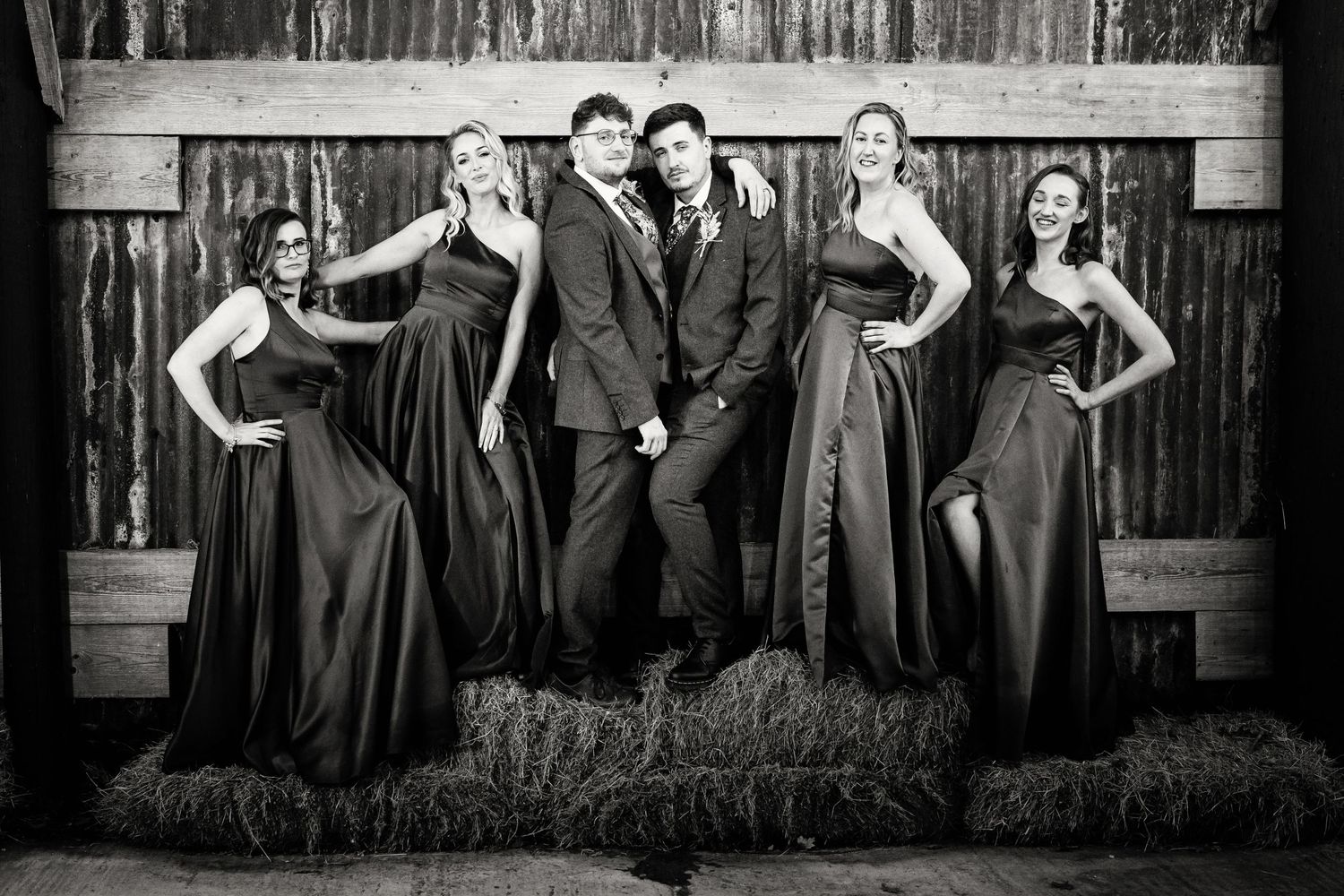 Black and white portrait of wedding party posed on hay bales in formal attire.