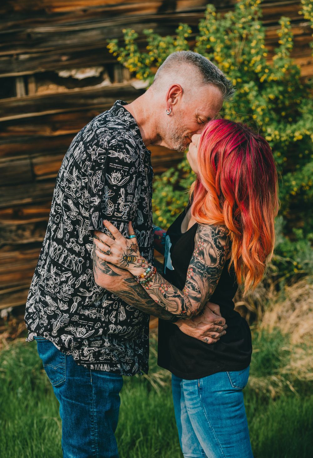 A couple with colorful hair and tattoos embrace against a rustic wooden wall backdrop.
