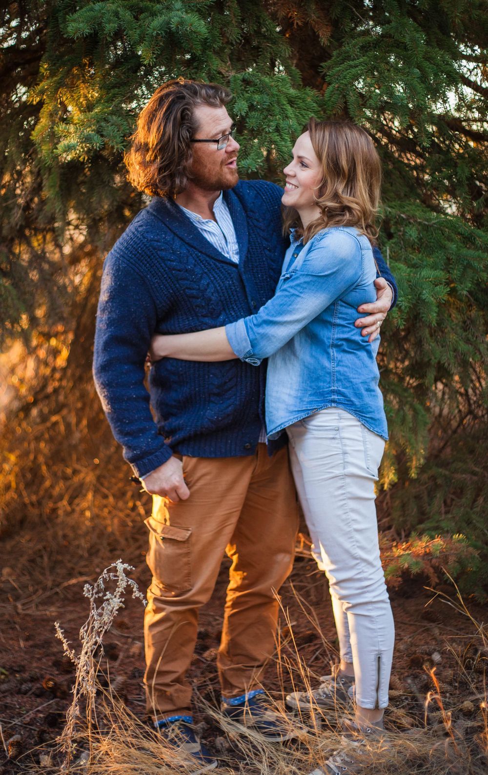A couple embraces outdoors against evergreen trees in warm sunset lighting.