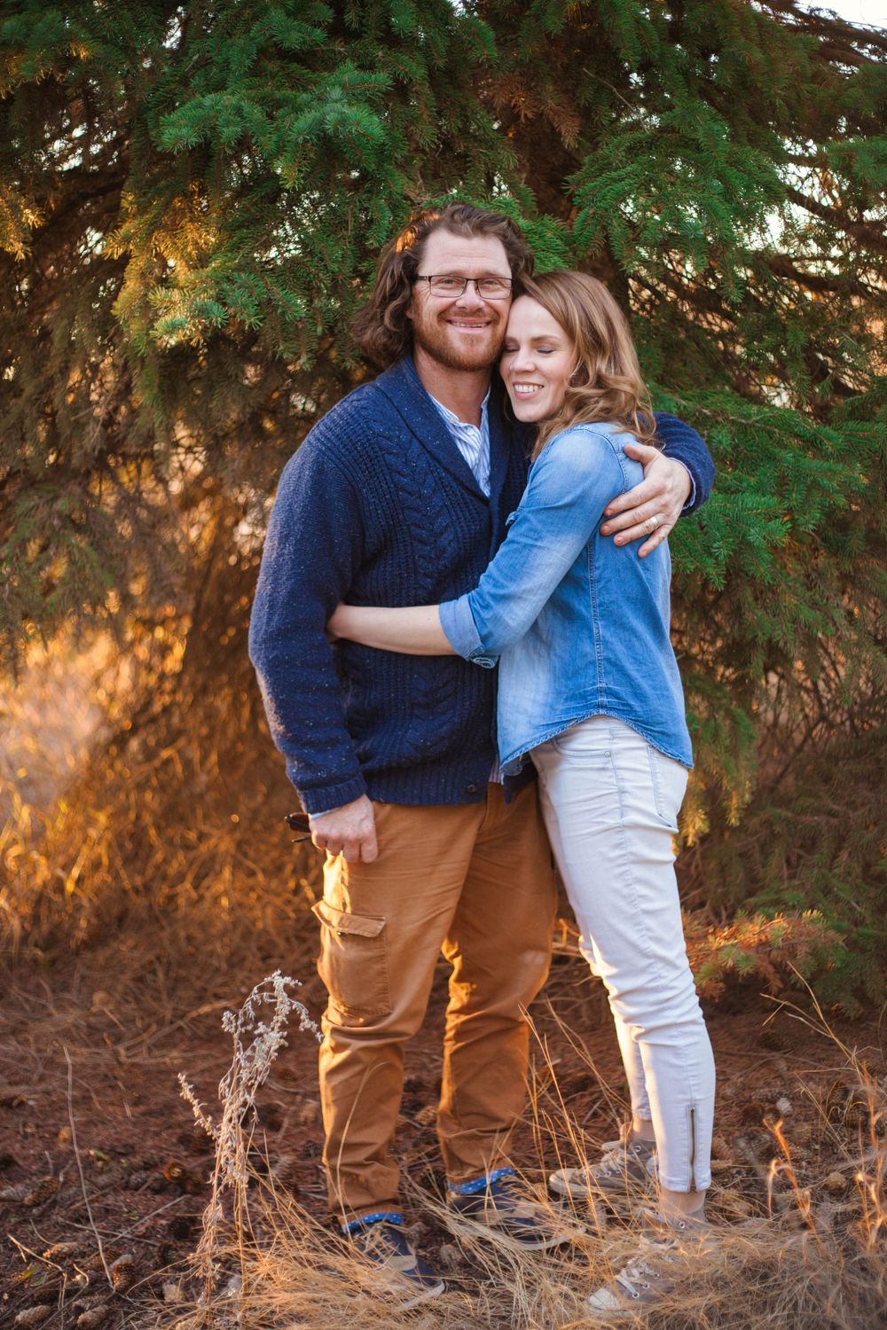 A romantic outdoor portrait of a couple in coordinated casual wear against pine trees.