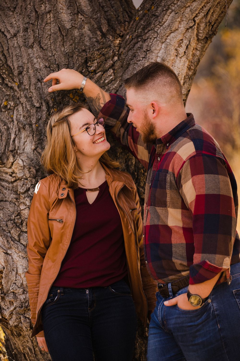 Romantic couple sharing tender moment against tree trunk in autumn setting with plaid shirt and leather jacket.