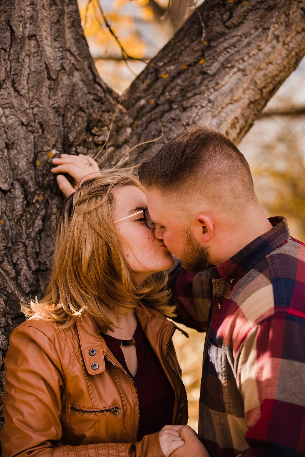 Intimate fall photoshoot of couple kissing by tree trunk in warm autumn lighting.