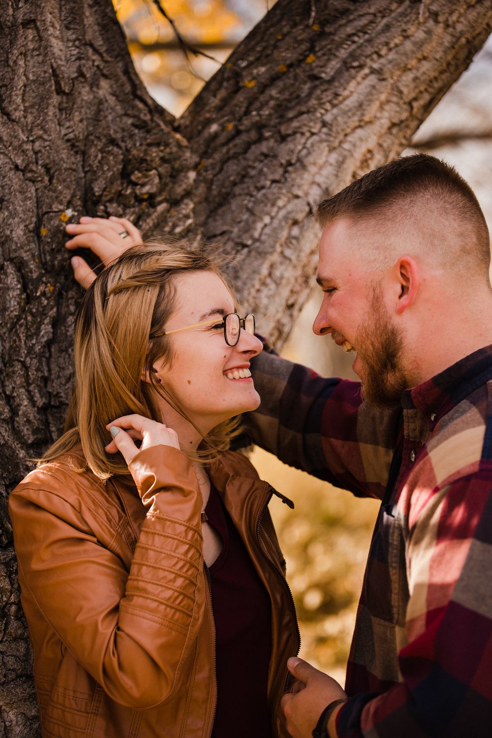 Playful couple laughing together against rustic tree trunk during autumn photoshoot.