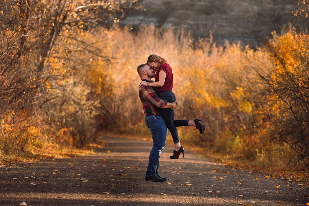 Playful autumn engagement photo with couple embracing on dirt path surrounded by fall foliage.