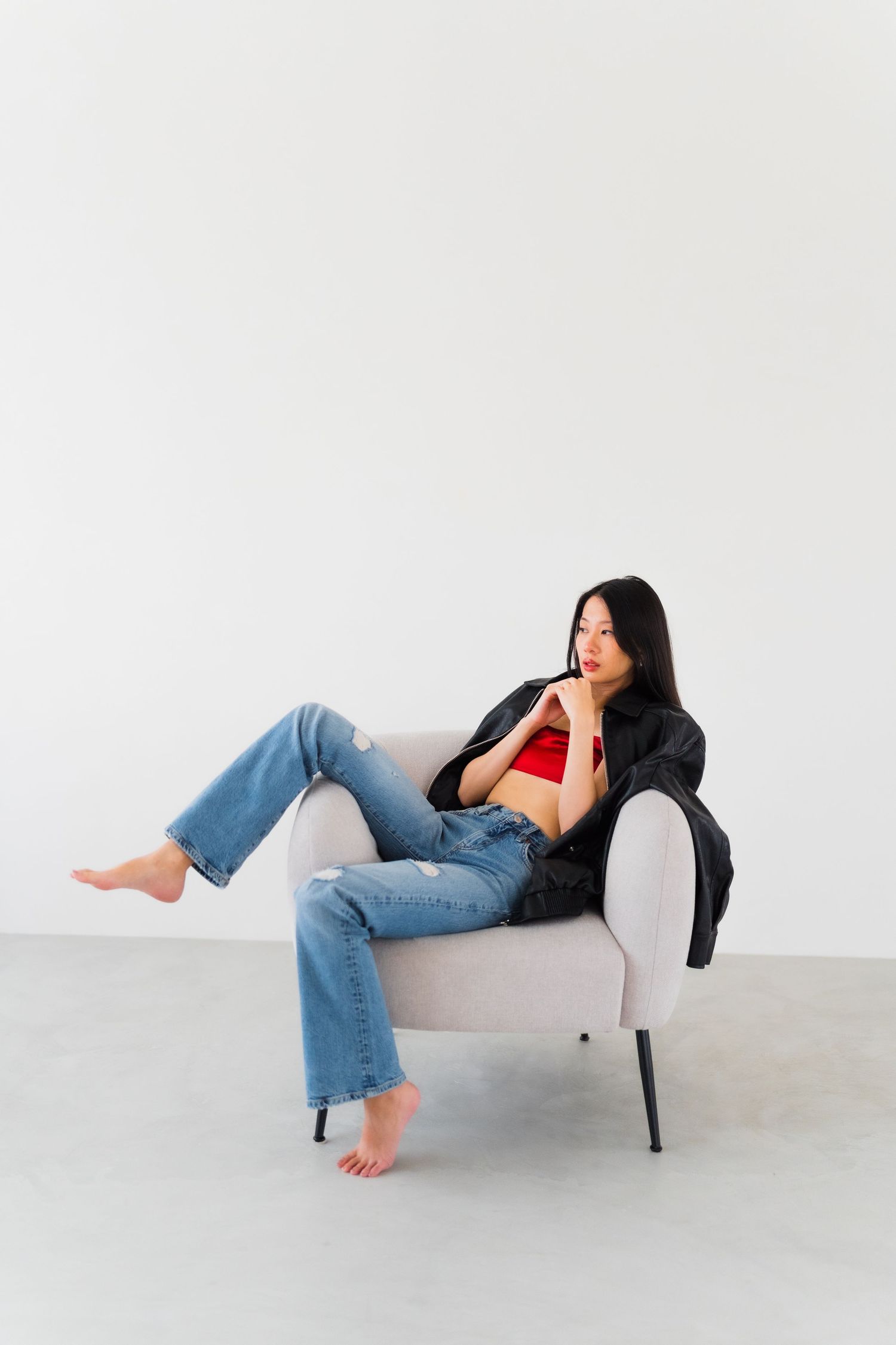 Woman relaxing barefoot on grey armchair against white wall wearing denim and black jacket.