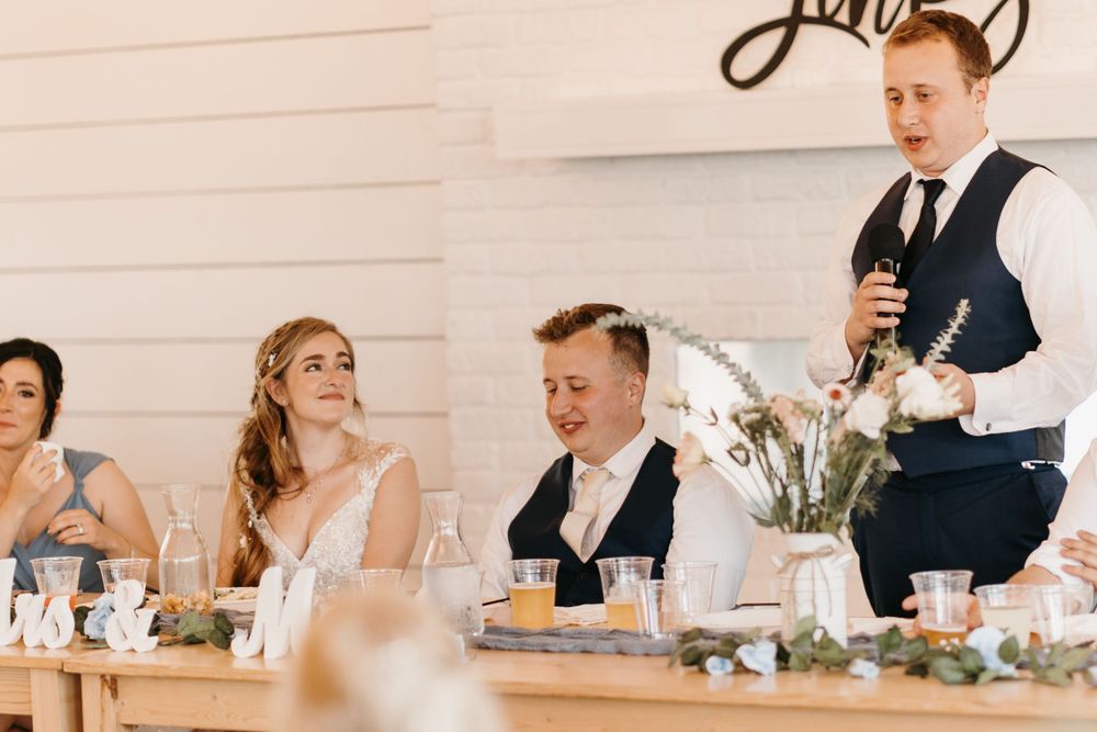 Groom gives a speech at wedding reception as bride and guests smile at a rustic white barn venue.