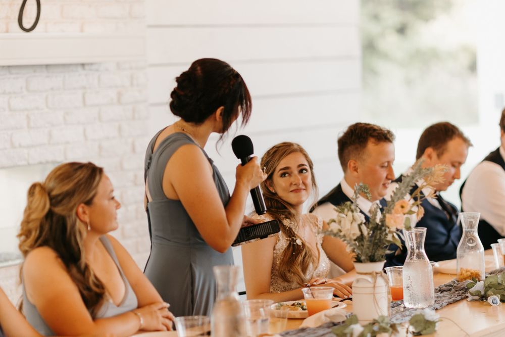 Bridesmaid speaks into microphone as bride and guests smile during wedding reception toast.