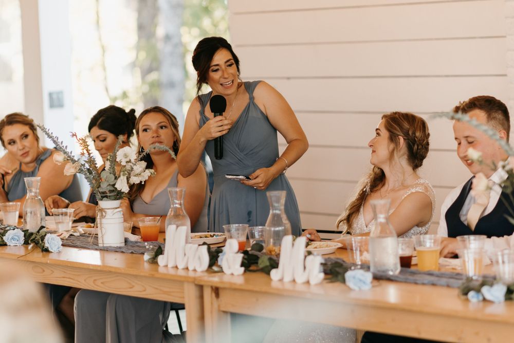 Smiling bridesmaid gives toast with microphone as wedding party laughs at decorated reception table.