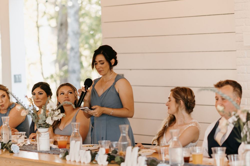 Bridesmaid in slate blue dress delivers heartfelt speech at elegant wedding reception dinner.