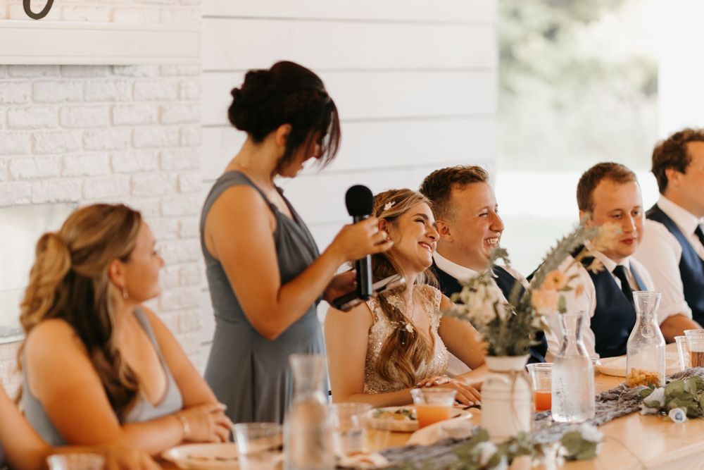 Wedding party laughs joyfully during bridesmaid toast at floral-decorated reception dinner table.