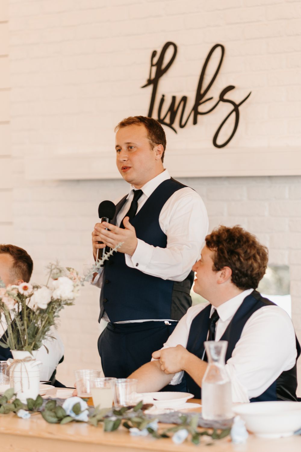 Groomsman delivers wedding toast with microphone beneath decorative script sign at reception.